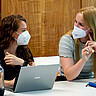 Das Symbolbild zeigt zwei Studentinnen. Sie sitzen in einem Seminarraum der Hochschule. Vor ihnen steht jeweils ein Laptop. Sie schauen sich freundlich an. Beide tragen corona-bedingt einen Mund-Nase-Schutz.