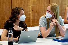 Das Symbolbild zeigt zwei Studentinnen. Sie sitzen in einem Seminarraum der Hochschule. Vor ihnen steht jeweils ein Laptop. Sie schauen sich freundlich an. Beide tragen corona-bedingt einen Mund-Nase-Schutz.