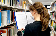 Auf dem Bild sieht man eine Frau von hinten. Sie steht in der Bibliothek und liest. Corona-bedingt trägt sie einen Mund-Nase-Schutz.