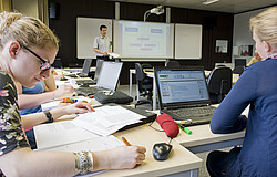 Das Bild zeigt Studierende an Tischen die Schreiben und vor ihren Laptops sitzen.