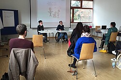 zwDas Bild zeigt die Rückenansicht von Menschen, die einem Vortrag von zwei Referenten zuhören.