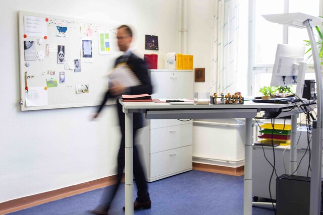  Ein Büro im Altbau der Pädagogischen Hochschule Heidelberg  mit Steh-Sitz-Arbeitstisch im Vordergrund. Ein Mann läuft hinter dem Schreibtisch hervor; er ist lediglich schemenhaft sichtbar. 