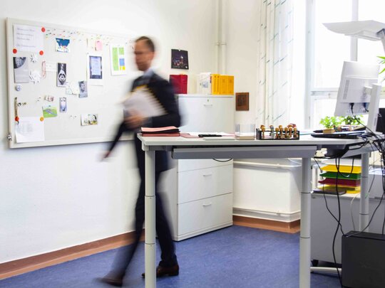 Ein Büro mit Steh-Sitz-Arbeitstisch im Vordergrund. Ein Mann läuft hinter dem Schreibtisch hervor; er ist lediglich schemenhaft sichtbar.