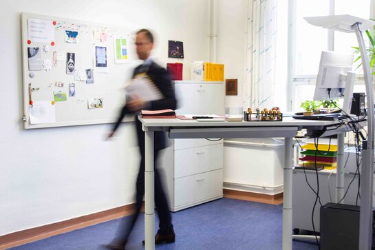Ein Büro mit Steh-Sitz-Arbeitstisch im Vordergrund. Ein Mann läuft hinter dem Schreibtisch hervor; er ist lediglich schemenhaft sichtbar.