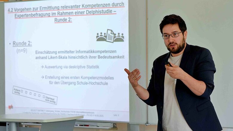 Ein junger Mann spricht deutlich mit den Händen. Hinter ihm sieht man ein Whiteboard.