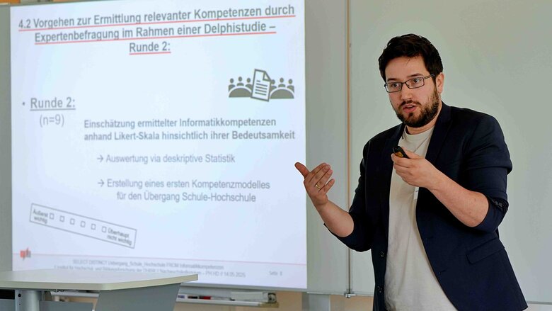 Ein junger Mann spricht deutlich mit den Händen. Hinter ihm sieht man ein Whiteboard.
