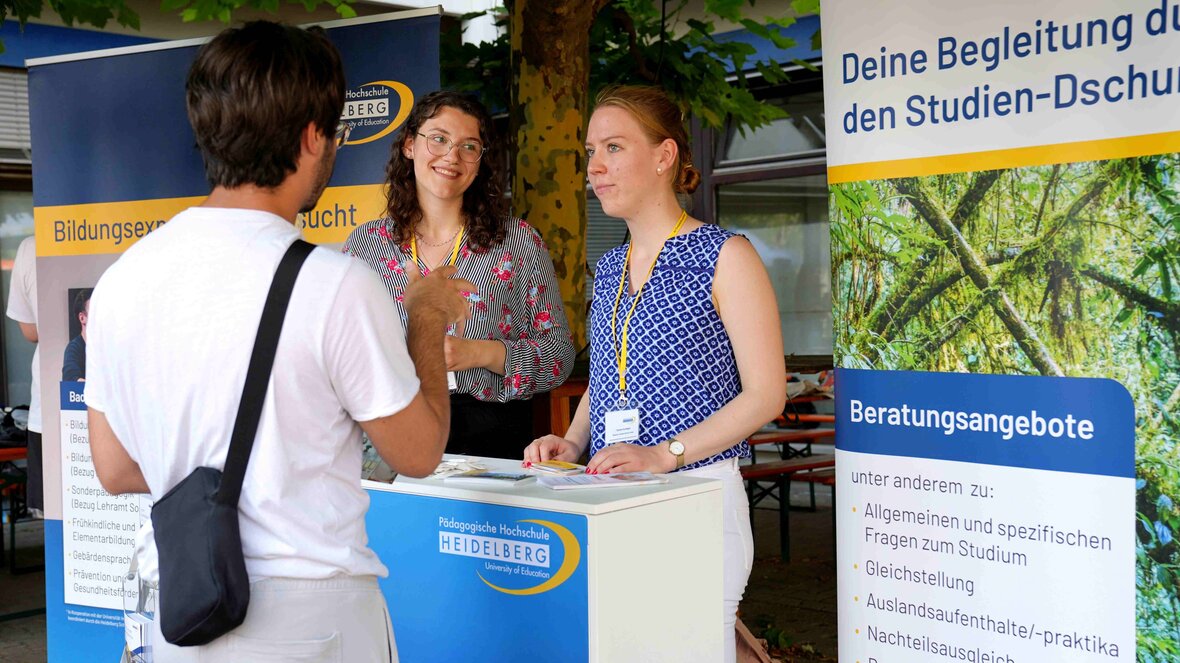 Ein junger Mann wird am Infostand der PHHD von zwei studentischen Tutorinnen beraten. 