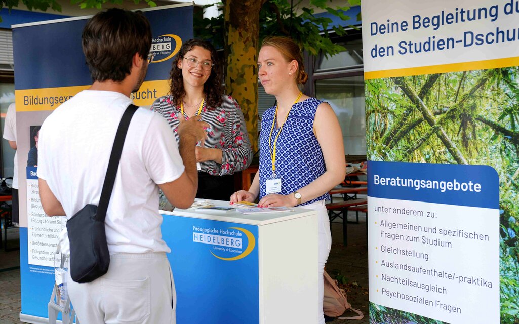 Ein junger Mann wird am Infostand der PHHD von zwei studentischen Tutorinnen beraten. 