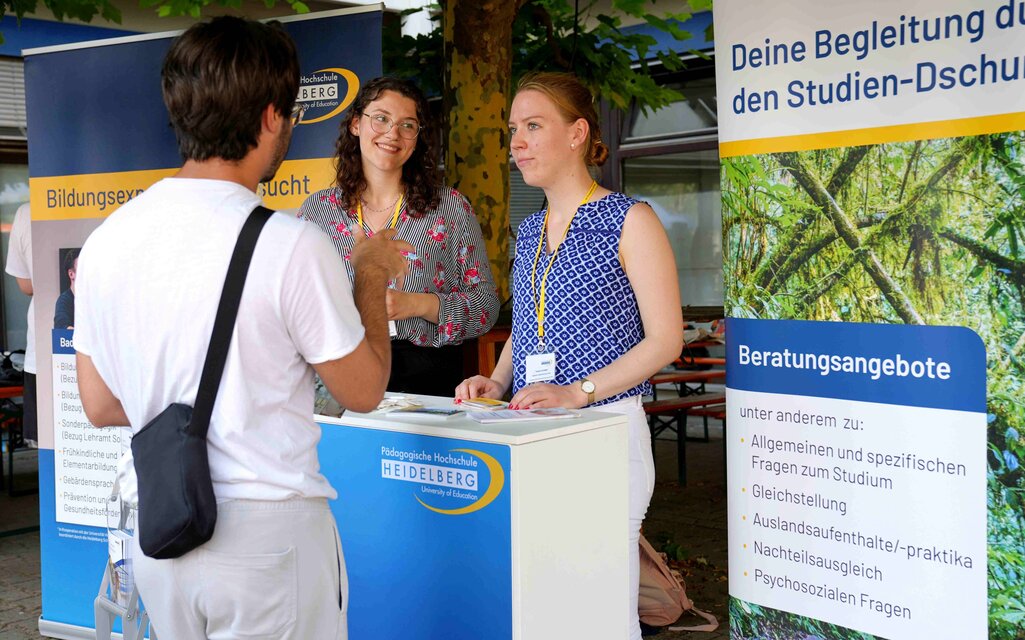 Ein junger Mann wird am Infostand der PHHD von zwei studentischen Tutorinnen beraten. 