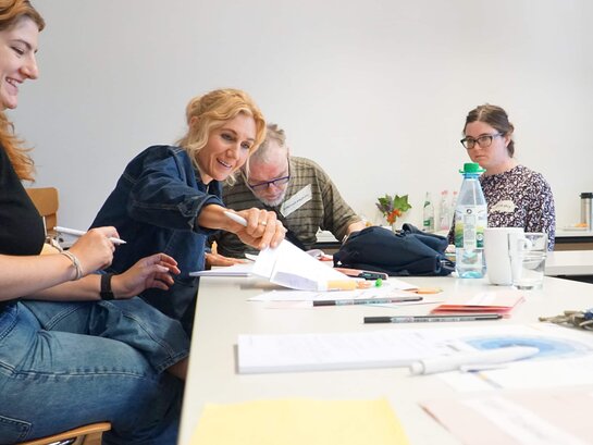 Das Bild zeigt Teilnehmende des Workshops, die an einem Tisch mit vielen Papieren sitzen. Eine Frau greift ein Stück Papier.