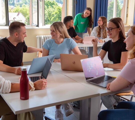 Eine Gruppe Studierender sitzt an einem Tisch und diskutiert. Im Hintergrund sieht man drei Student:innen, ebenfalls im Gespräch.