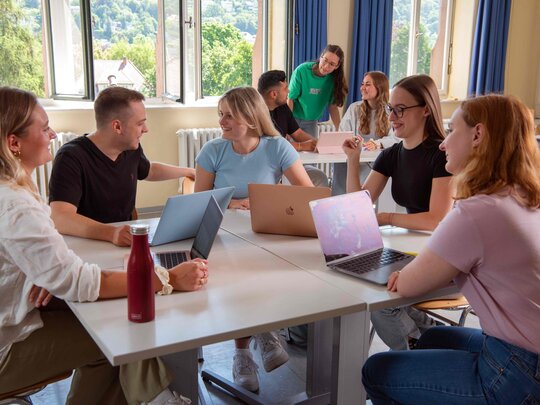 Eine Gruppe Studierender sitzt an einem Tisch und diskutiert. Im Hintergrund sieht man drei Student:innen, ebenfalls im Gespräch.