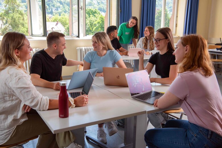 Eine Gruppe Studierender sitzt an einem Tisch und diskutiert. Im Hintergrund sieht man drei Student:innen, ebenfalls im Gespräch.