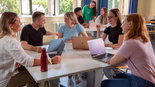 Eine Gruppe Studierender sitzt an einem Tisch und diskutiert. Im Hintergrund sieht man drei Student:innen, ebenfalls im Gespräch.