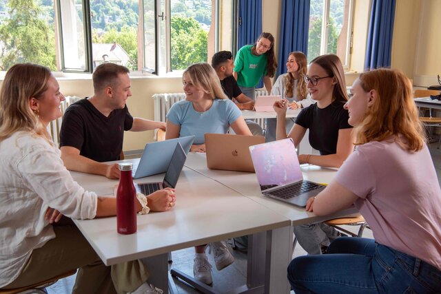 Eine Gruppe Studierender sitzt an einem Tisch und diskutiert. Im Hintergrund sieht man drei Student:innen, ebenfalls im Gespräch.
