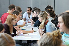 Das Bild zeigt sich an Tischen gegenübersitzende Studierende, die Unterhaltungen führen.