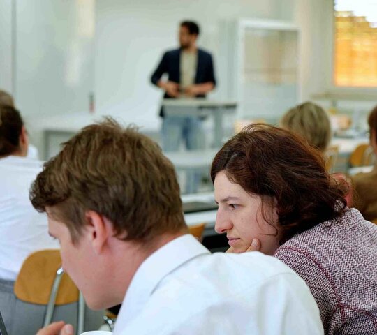 Ein Mann präsentiert vorne im Seminarraum, Studierende machen sich Notizen.