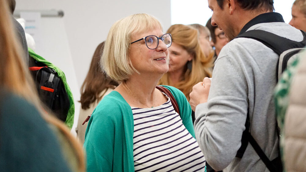 Manuela Welzel-Breuer im Gespräch mit jemanden. Im Hintergrund sieht man unscharf weitere Mitglieder der Hochschule.
