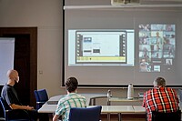 Auf dem Bild sieht man Till Krüger von der Seite und Michael Gänßmantel sowie Helmuth Pflantzer von hinten. Sie schauen auf eine zoom-Präsentation, die groß auf der Leinwand zu sehen ist. Der Hintergrund ist verschwommen. Das Bild wurde im Senatssaal der Hochschule aufgenommen. 