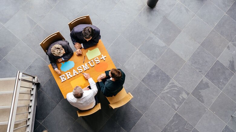 Aus der Vogelperspektive sehen wir vier Personen an einem Informationstisch zu Angeboten von Erasmus+ sitzen.