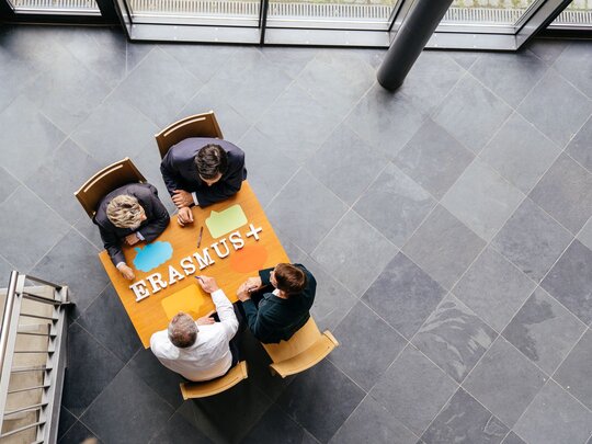 Aus der Vogelperspektive sehen wir vier Personen an einem Informationstisch zu Angeboten von Erasmus+ sitzen.