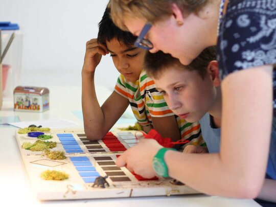 Das Foto zeigt eine Lehrende die zwei Kindern etwas in einem Buch zeigt.