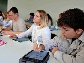 Studierende sitzen an einem Tisch und schreiben auf Tablets.