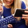 Das Symbolbild fokussiert auf ein Handy. Im Hintergrund sieht man unscharf ein kleines Mädchen mit seiner Mutter, die auf das Handy schauen. 