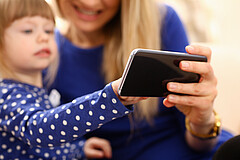 Das Symbolbild fokussiert auf ein Handy. Im Hintergrund sieht man unscharf ein kleines Mädchen mit seiner Mutter, die auf das Handy schauen. 