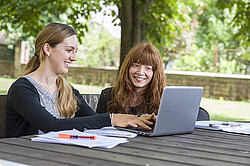 Das Bild zeigt zwei lachende Studierende die zusammen an einem Laptop arbeiten.