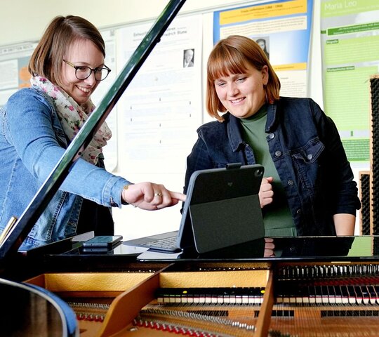 Teilnehmende stehen vor einem Klavier, darauf ein Tablet, eine Person tippt auf das Tablet, Lachen
