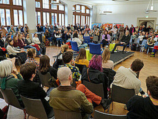 Das Bild zeigt die Aula der PH Heidelberg. Viele Menschen sind im Raum, in der Mitte ist eine etwas erhöhte Bühne mit Plätzen für die Diskussionsteilnehmenden. 