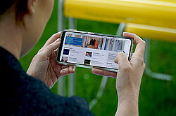 Auf dem Bild sieht man eine Frau von schräg hinten. Sie hält ein Handy quer in der Hand. Auf dem Handy sieht man die Startseite der Website der Hochschule. Handy in der Hand