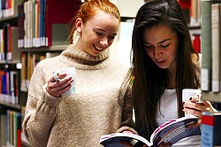 Das Bild zeigt zwei Frauen nebeneinander stehen. Die eine Frau hält ein Buch in der Hand, und die andere Frau ein Tasse. Beiden schauen in das Buch. Im Hintergrund sind Bücherregale zu erkennen.