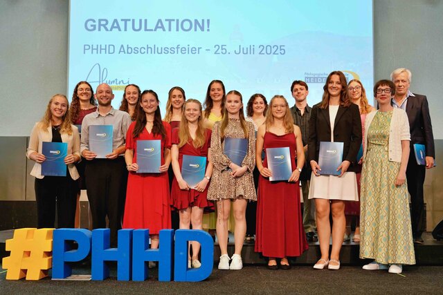 Zu sehen sind Alumni, die auf einer Bühne stehen und ihre Bestehensbriefe in PHHD-Mappen hochhalten. Gemeinsam mit Rektorin Vach & Prorektor Dammer (a.D.), welche ihnen die Mappen überreicht haben.