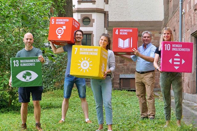 Drei Männer und zwei Frauen stehen auf der Wiese im Altbau und halten Würfel mit SDG-Zielen hoch. Diese spielen für nachhaltige Lehre eine bedeutende Rolle.