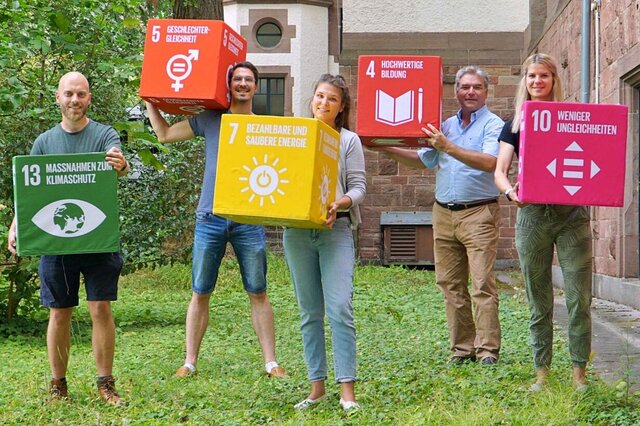 Drei Männer und zwei Frauen stehen auf der Wiese neben dem Altbau. Und halten verschiedene SDG-Würfel hoch.