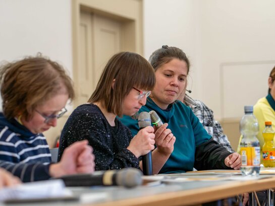 Qualifizierungsteilnehmerin Susann Bensch sitzt an einem Tisch und spricht in ein Mikrofon, das Noemi Heiser hält. Im Vordergrund ist Louisa Kabbe, im Hintergund Sarah Maier. 