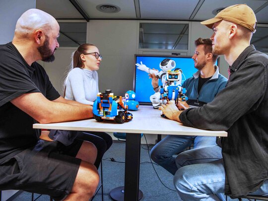 Vier Personen sitzen an einem Tisch auf dem mehrere Roboter stehen.