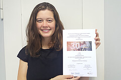 Das Bild zeigt die Studentin Lara-Dora Ellerbrock. Sie hält die Urkunde mit der Auszeichnung hoch und lächelt in die Kamera.