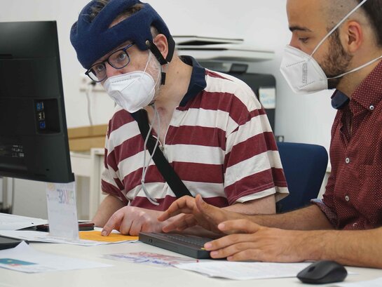 Das Bild zeigt zwei Personen, die gemeinsam vor einem Computer-Bildschirm sitzen und arbeiten.