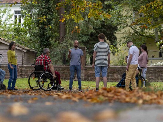 Das Bild zeigt eine Gruppe von Menschen, die im Innenhof der PH Heidelberg im Halbkreis stehen bzw. im Rollstuhl sitzen. Sie machen Entspannungsübungen. Im Vordergund ist buntes Herbstlaub.