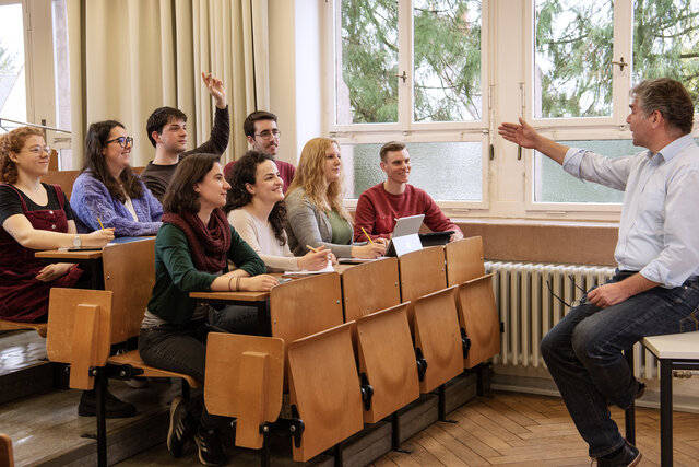 Ein Dozent sitzt vo einer Gruppe Studierender und zeigt mit einer Hand auf eine Person in der Hörerschaft, die ihre Hand hebt.