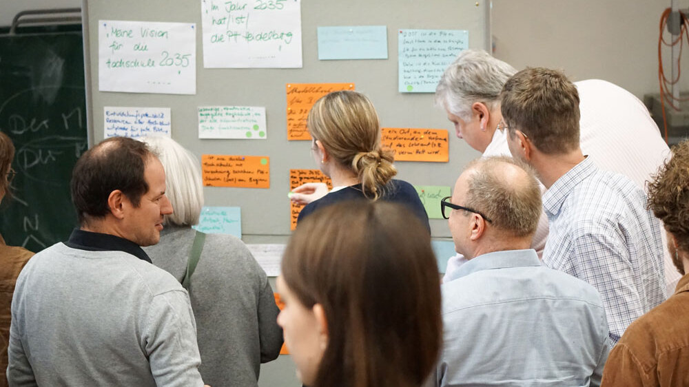 Mehrere Menschen stehen vor einer Pinnwand. Einige blicken auf die Wand. Andere sprechen miteinander.