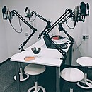 Auf dem Bild sieht man einen Raum mit einem Tisch, vier Hocker und vier Mikrofonarme - das ist Podcaststudio.