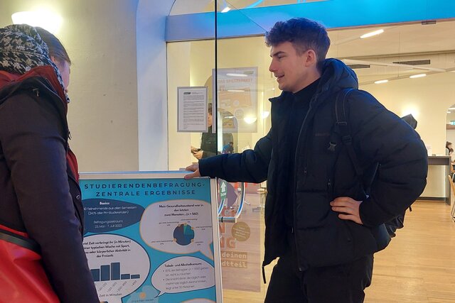 Zwei Studierende stehen vor einem Plakat, auf dem die Ergebnisse der Studierendenbefragung von PHeelGood zu sehen sind. Im Hintergrund ist die Mensa im Altbau der Pädagogischen Hochschule Heidelberg zu sehen. 