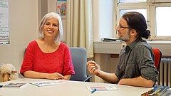 Frauke Janz schaut lächelnd direkt in die Kamera. Wolfgang Schultz schaut sie an und spricht deutlich mit den Händen-