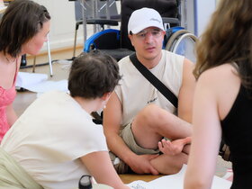Qualifizierungsteilnehmer Mehmet Can Friedel sitzt auf dem Boden und diskutiert mit Studierenden über Lenrbarrieren. Eine Studentin schreibt auf ein Flip-Chart-Papier, das auf dem Boden liegt.