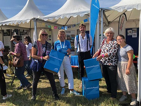 Zu sehen sind fünf Leute auf einem Festgelände mit blauen Logos.