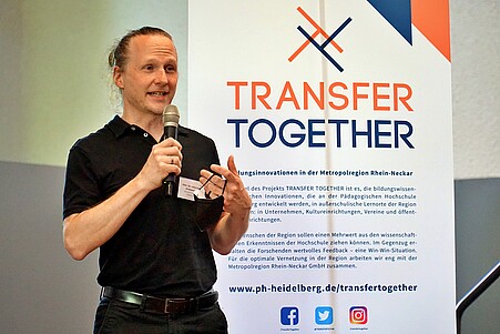 Zu sehen ist ein Mann vor einem Roll-Up mit dem Titel "Transfer Together"